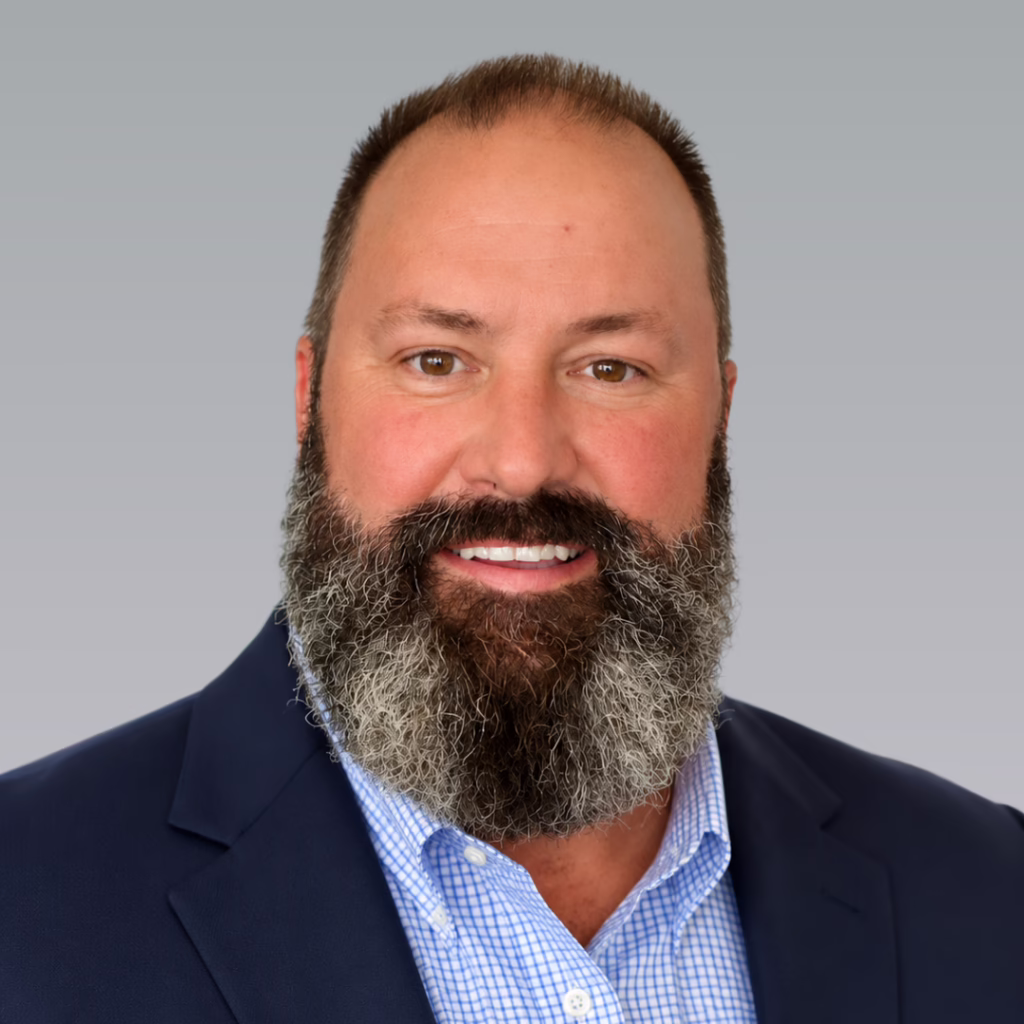 Professional head-and-shoulders portrait of a smiling man with a full beard, wearing a navy blazer and blue checkered shirt against a gray gradient background.